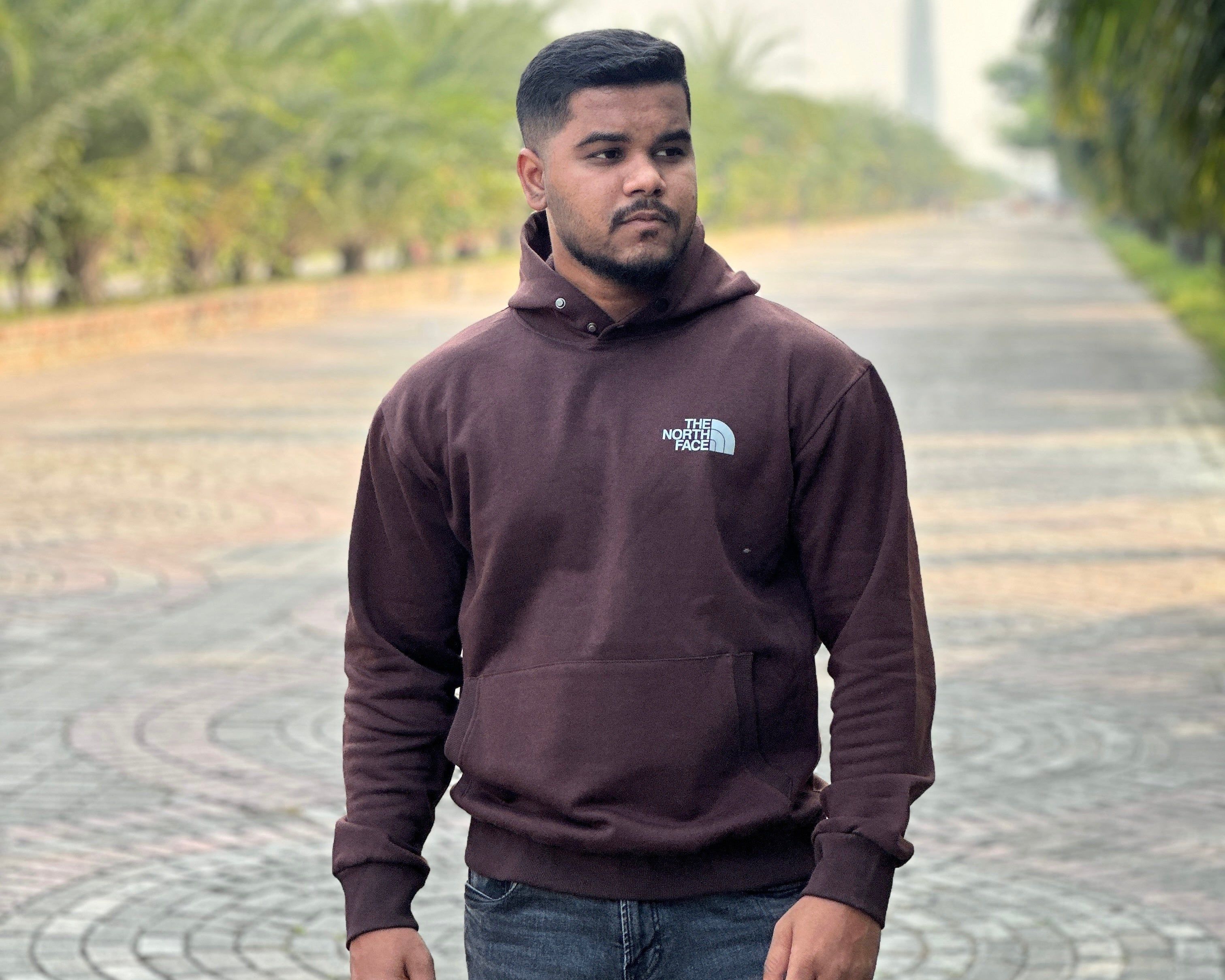 Man wearing a brown hoodie with a logo standing on a paved path with greenery in the background